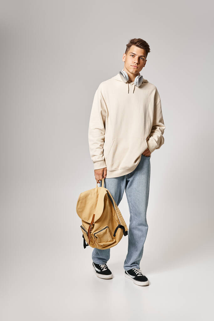 pohledný student ve sluchátkách a ležérní oblečení chůze s batohem na šedém pozadí - Fotografie, Obrázek