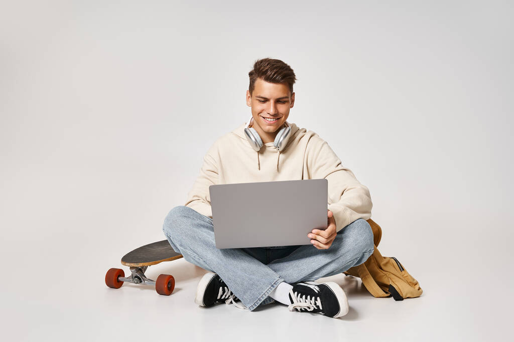 inteligentny student w sieci słuchawek do laptopa i siedzi z plecakiem i deskorolką - Zdjęcie, obraz