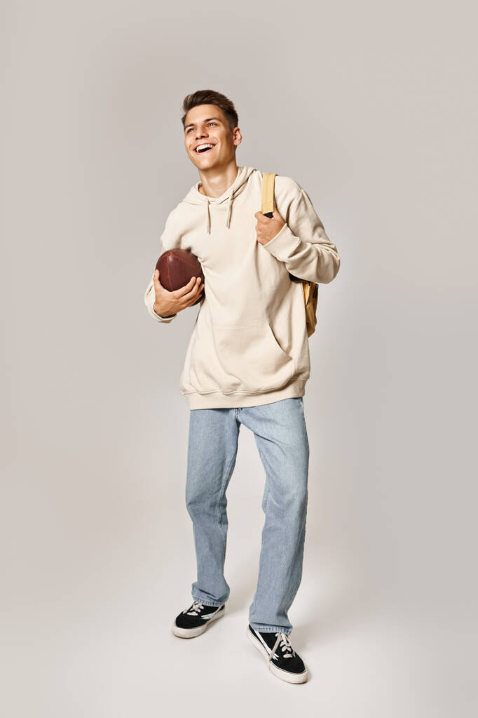 affascinante giovane studente in abito casual con zaino in cerca di e ridere con la palla da rugby - Foto, immagini