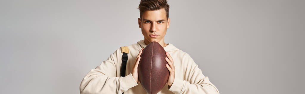 グレーの背景に手でラグビーボールを保持するバックパックで自信を持った学生の水平ショット - 写真・画像