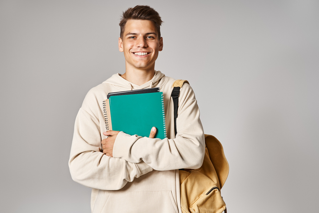 улыбающийся студент в возрасте 20 лет с рюкзаком, держащий заметки с руками на сером фоне - Фото, изображение