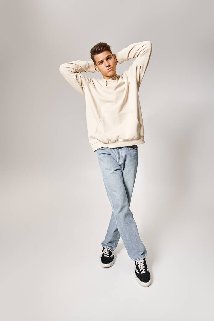 γοητευτικός νεαρός με casual ντύσιμο βάζει τα χέρια πίσω από το κεφάλι και στέκεται με σταυρωτά πόδια - Φωτογραφία, εικόνα