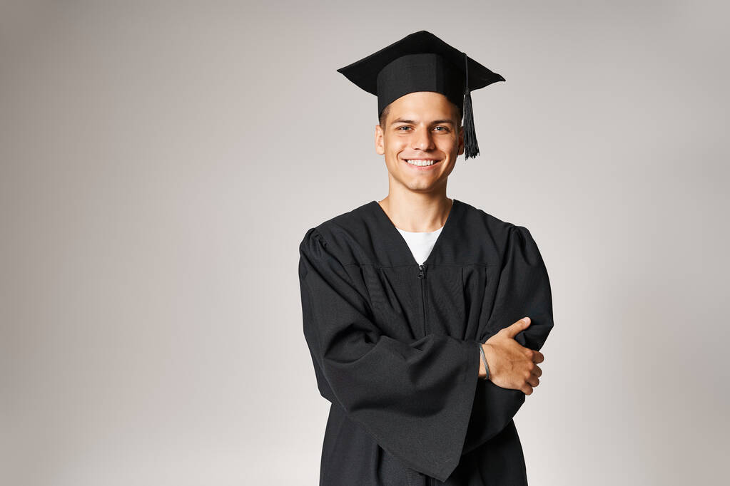 Gri gözlü, mezuniyet cüppeli ve şapkalı yakışıklı bir öğrenci açık arkaplanda kollarını kavuşturmuş. - Fotoğraf, Görsel