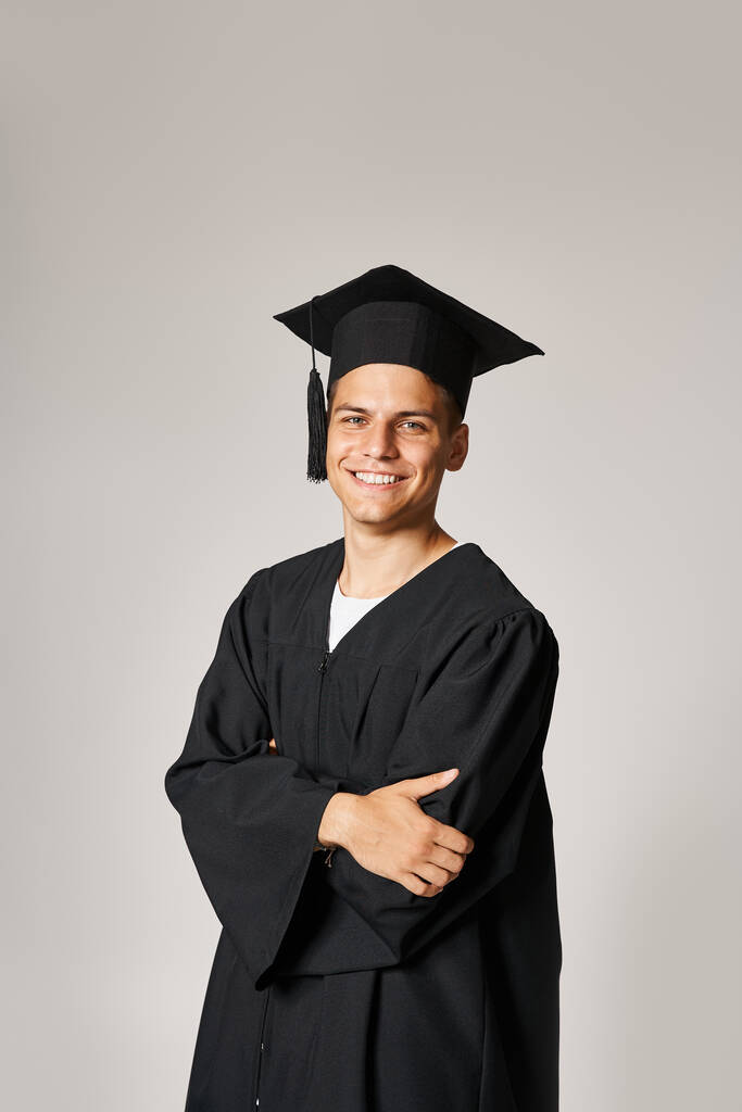 encantador estudiante en vestido de graduado y gorra con los brazos cruzados sonriendo y mirando a la cámara - Foto, Imagen