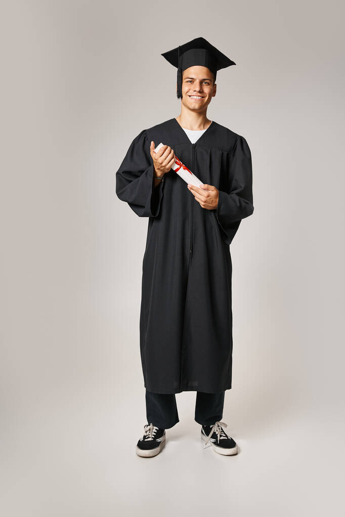 cheerful young student in graduate gown and cap holding to diploma with hands on grey background - Photo, Image