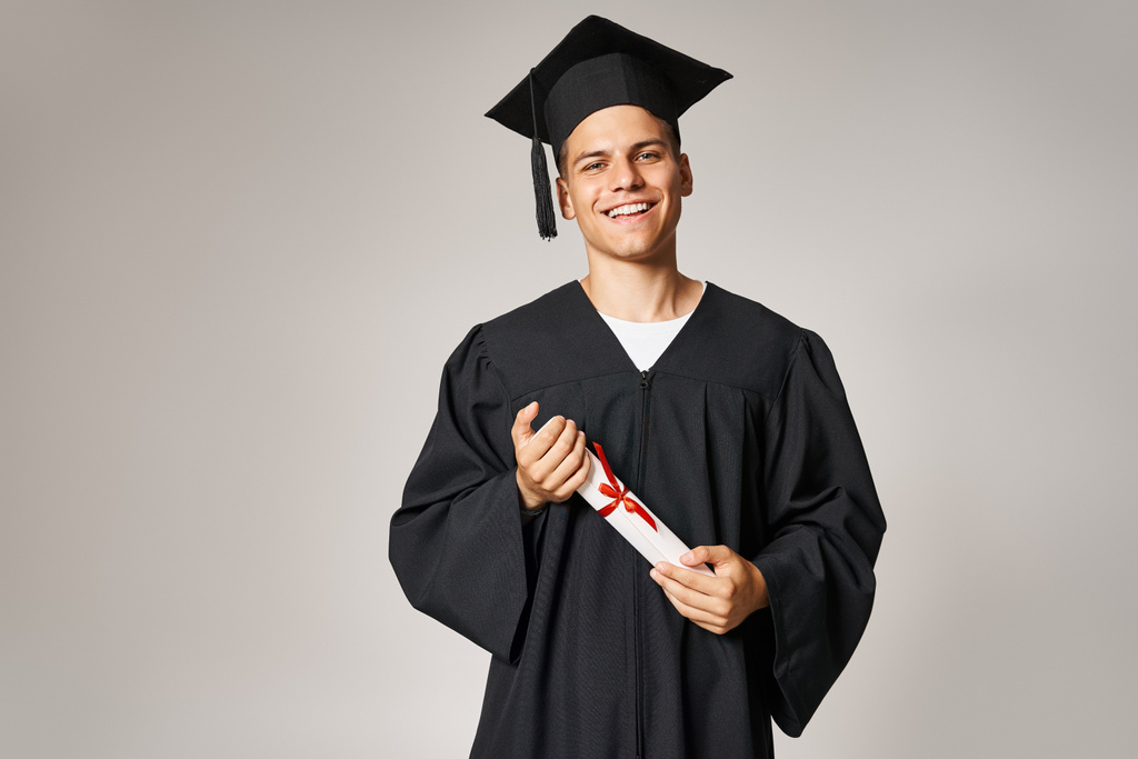 портрет молодого студента в аспірантурі та кепці, що тримається за диплом руками на сірому фоні - Фото, зображення