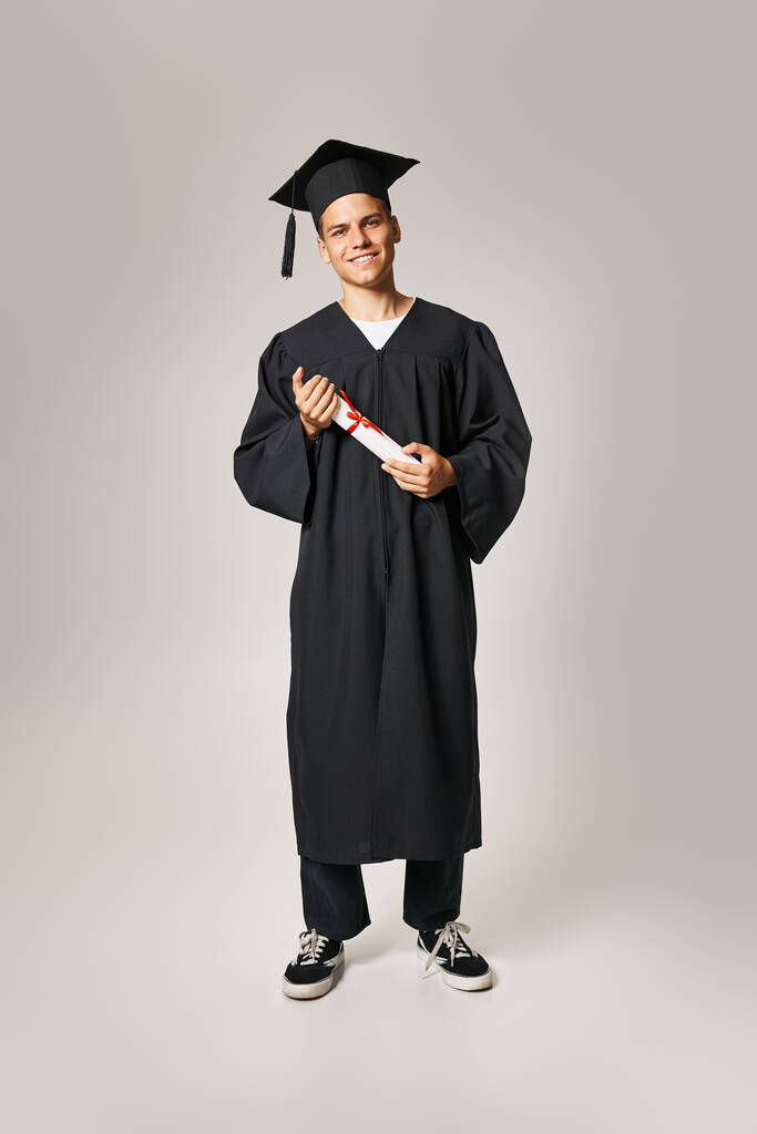 usmívající se student v absolventské šaty a čepice držící diplom s rukama na šedém pozadí - Fotografie, Obrázek