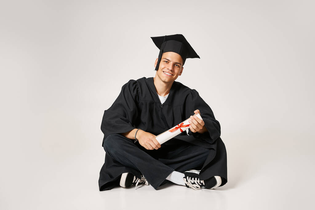bel homme en robe d'études supérieures et casquette assis et tenant au diplôme avec les mains sur fond gris - Photo, image