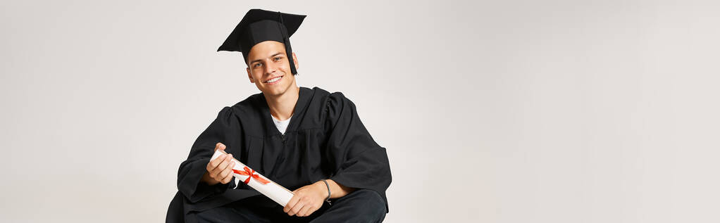 banner of happy man in graduate gown and cap sitting and holding to diploma with hands - Photo, Image