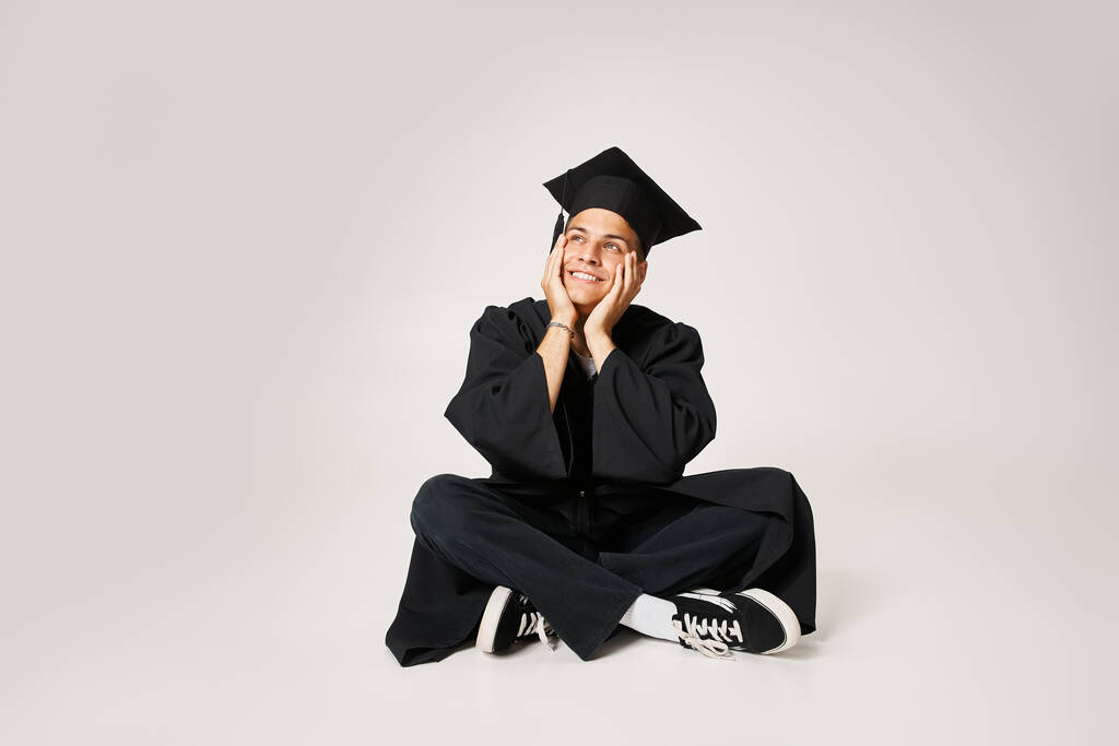 smiling charming guy in graduate outfit sitting and holding with hands to cheeks on grey background - Photo, Image