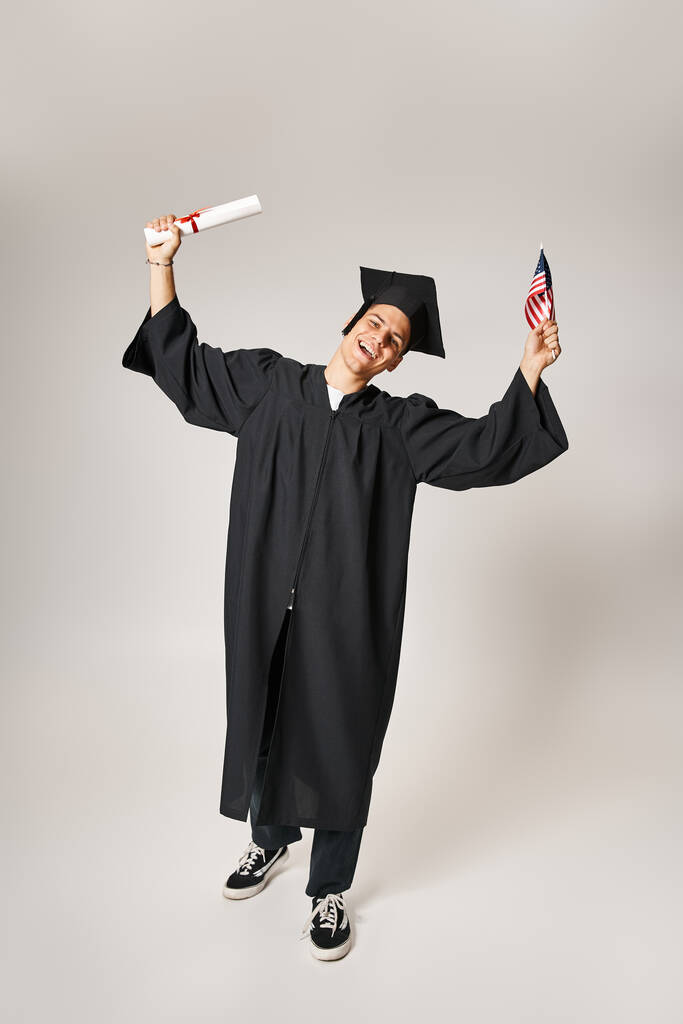 Američan mladý student v absolventské oblečení šťastný, že dokončil svá studia na šedém pozadí - Fotografie, Obrázek