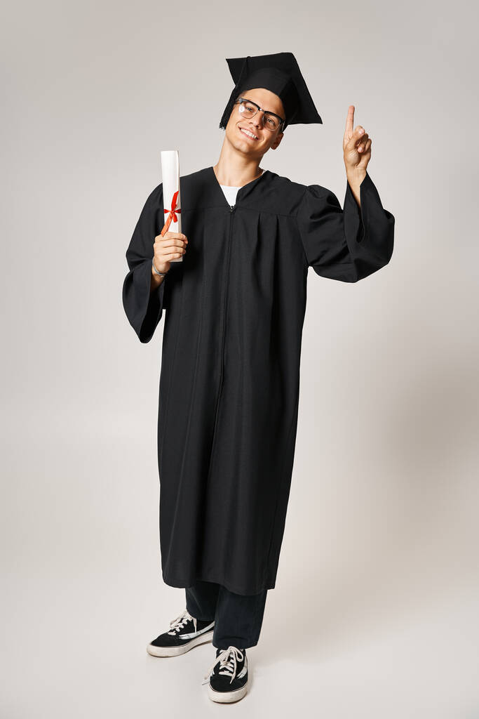 alegre joven en traje de graduado con gafas de visión apuntando con el dedo hacia arriba con diploma en la mano - Foto, imagen