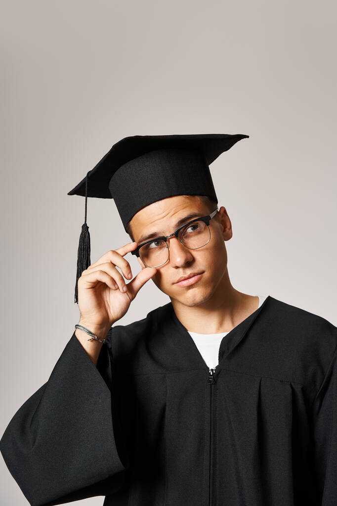 Charmanter junger Kerl im Diplom-Outfit, der die Hand an die Sehbrille legt und nach oben schaut - Foto, Bild