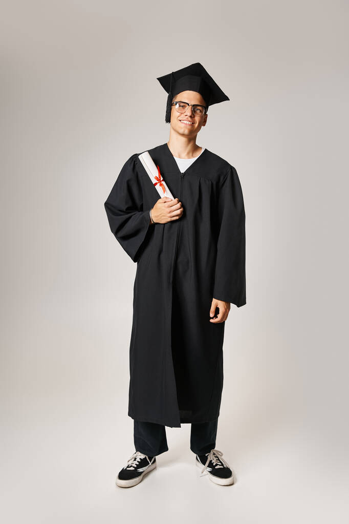 Mezuniyet kıyafetli ve görüş gözlüklü neşeli öğrenci elinde diplomayla ayakta duruyor. - Fotoğraf, Görsel