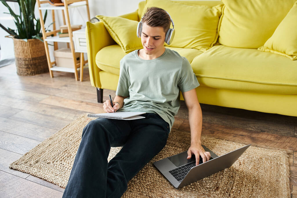 студент с наушниками на полу возле жёлтого дивана учится в ноутбуке и пишет заметки - Фото, изображение