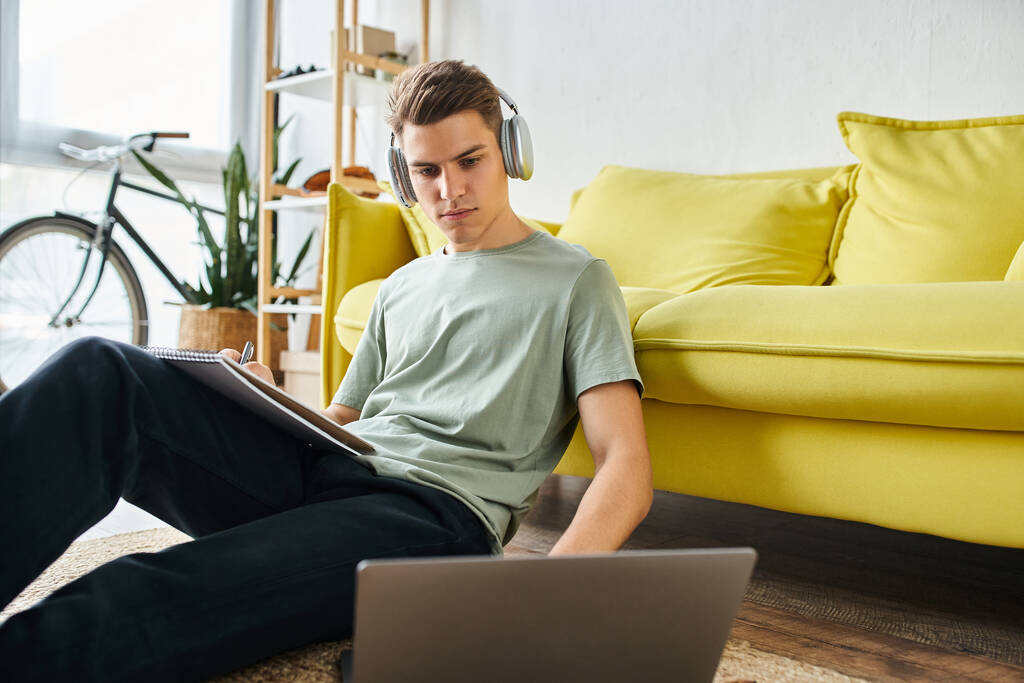 fokussierter junger Mann mit Kopfhörern auf dem Boden in der Nähe der gelben Couch, der in Laptop studiert und Notizen schreibt - Foto, Bild
