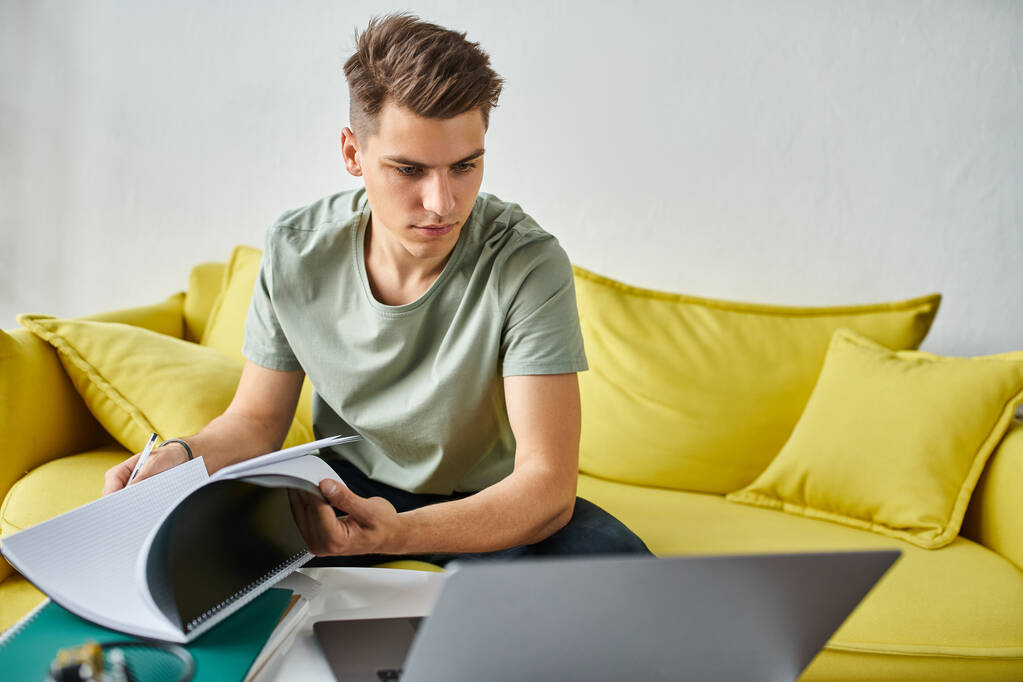 young man in yellow couch concentration doing coursework with notes and laptop on coffee table - Photo, Image