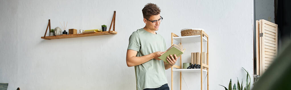 horizontal shot of young man with brown hair and vision glasses in living room reading book - Photo, Image