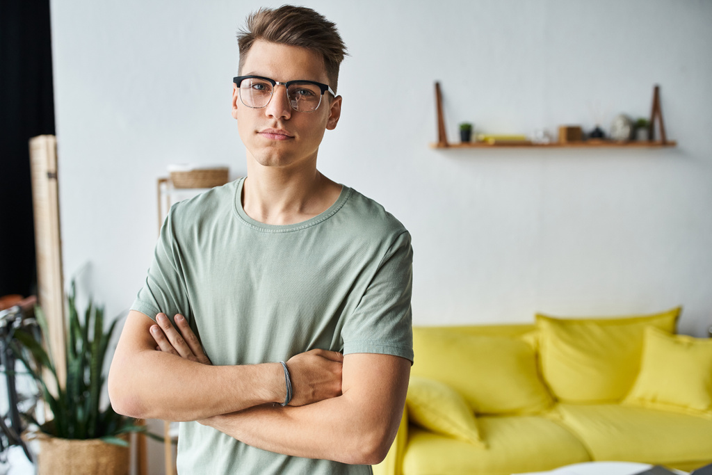 homem carismático em seus 20 anos com cabelos castanhos e óculos de visão na sala de estar braços cruzados dobráveis - Foto, Imagem