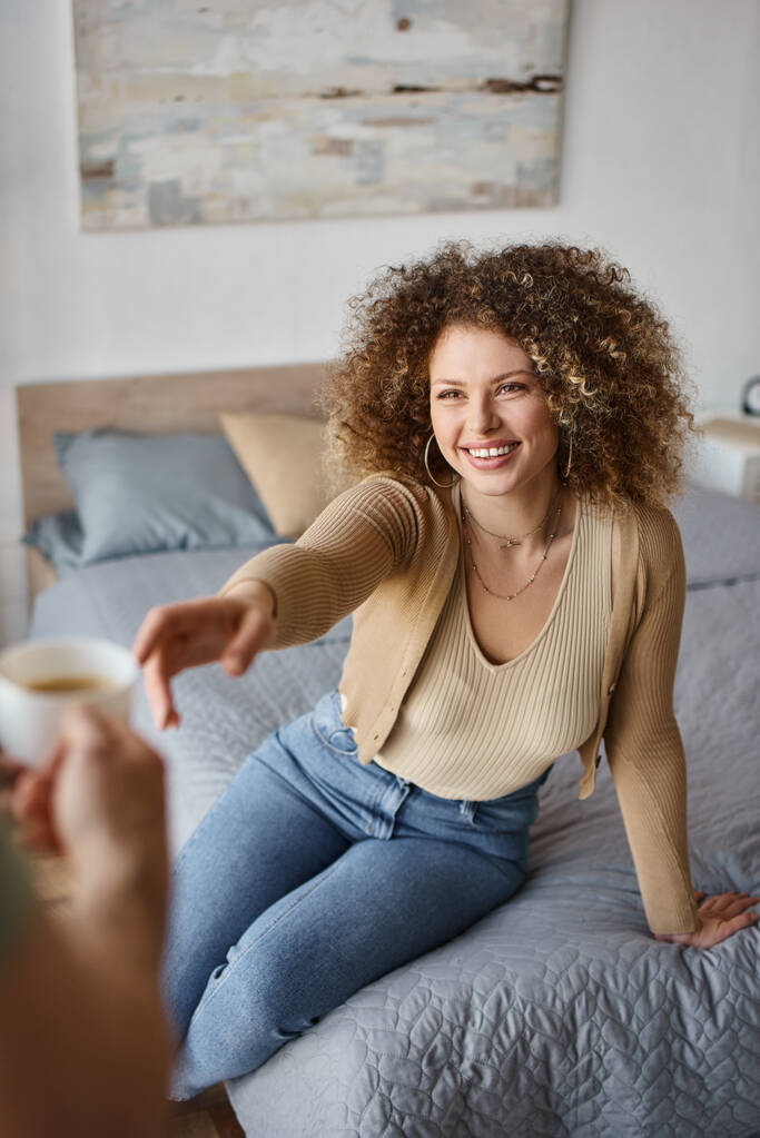 Kudrnatá žena usmívající se žena s šálkem kávy přítele ruku, pečující a užívající si čas doma - Fotografie, Obrázek