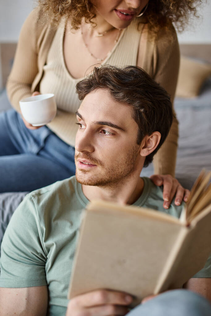Relaxed brunette man holding book and looking away, hugged by his girlfriend, morning couple time - Photo, Image