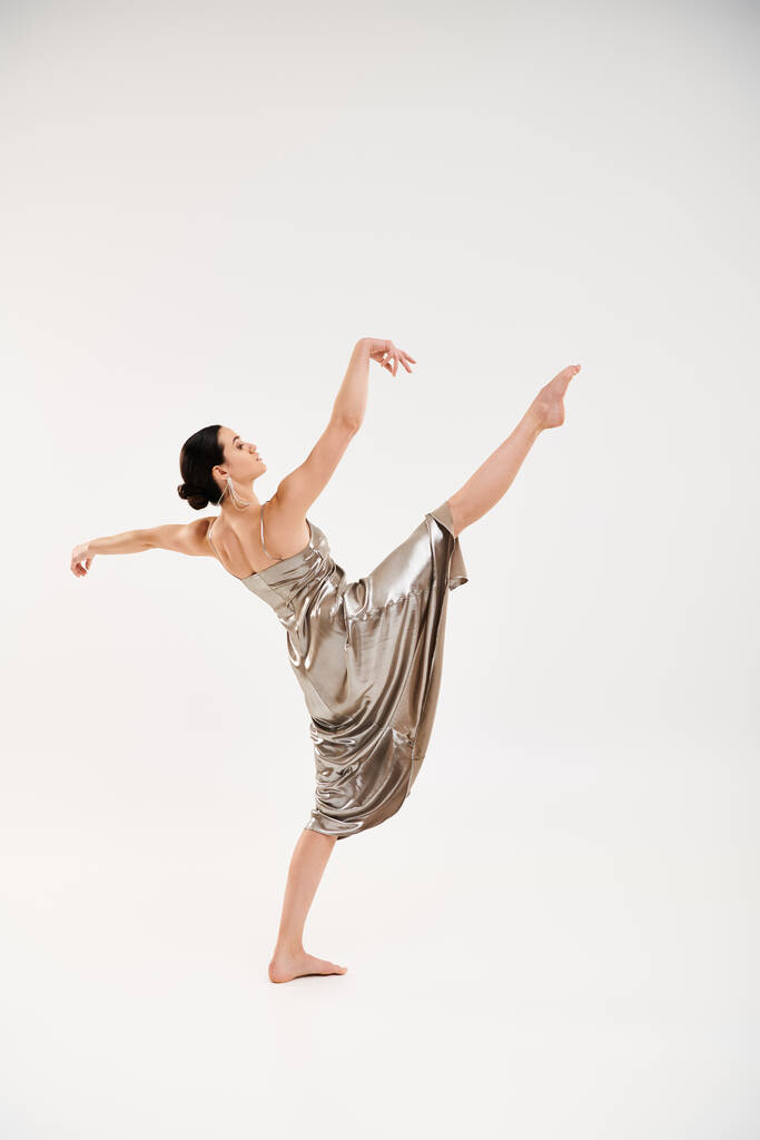 Egy kecses fiatal nő hosszú és fényes ezüst ruhában elegánsan táncol egy stúdióban, fehér háttérrel.. - Fotó, kép