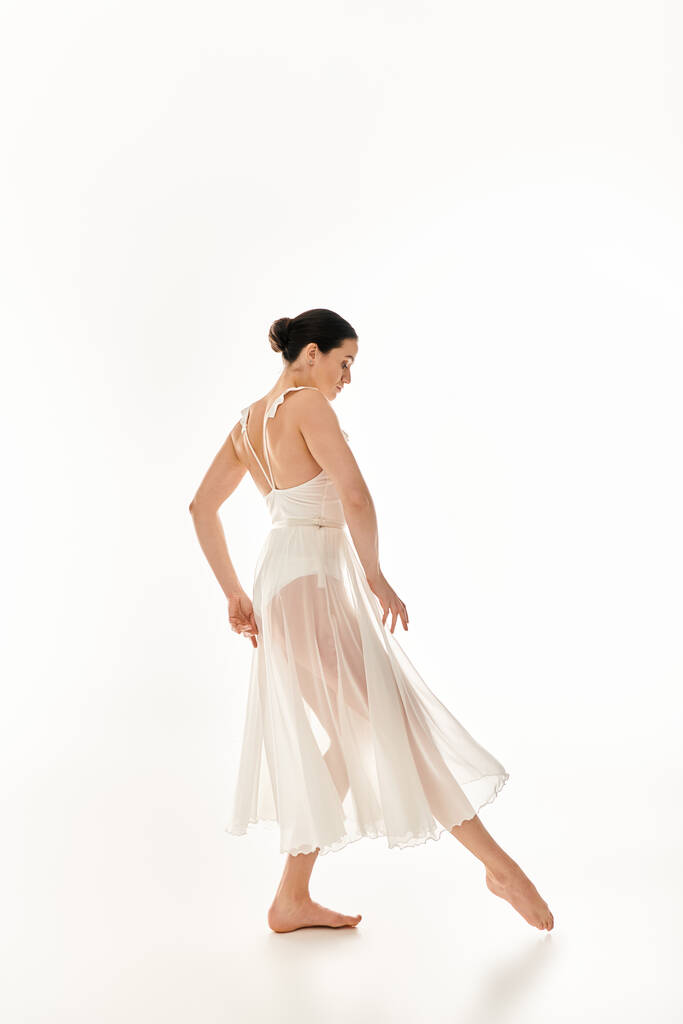 Een jonge vrouw in een vloeiende witte jurk danst sierlijk in een studio tegen een witte achtergrond. - Foto, afbeelding