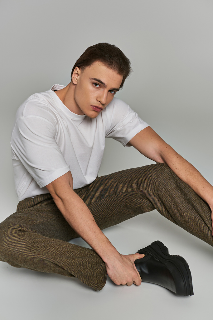 atractivo joven pulido en ropa elegante sentado sobre fondo gris y mirando a la cámara - Foto, Imagen