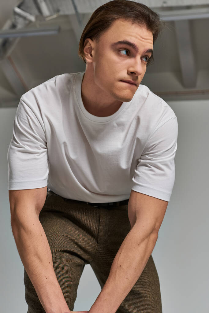 aantrekkelijke jongeman in t-shirt en bruine broek poseren aantrekkelijk op grijze achtergrond en kijken weg - Foto, afbeelding