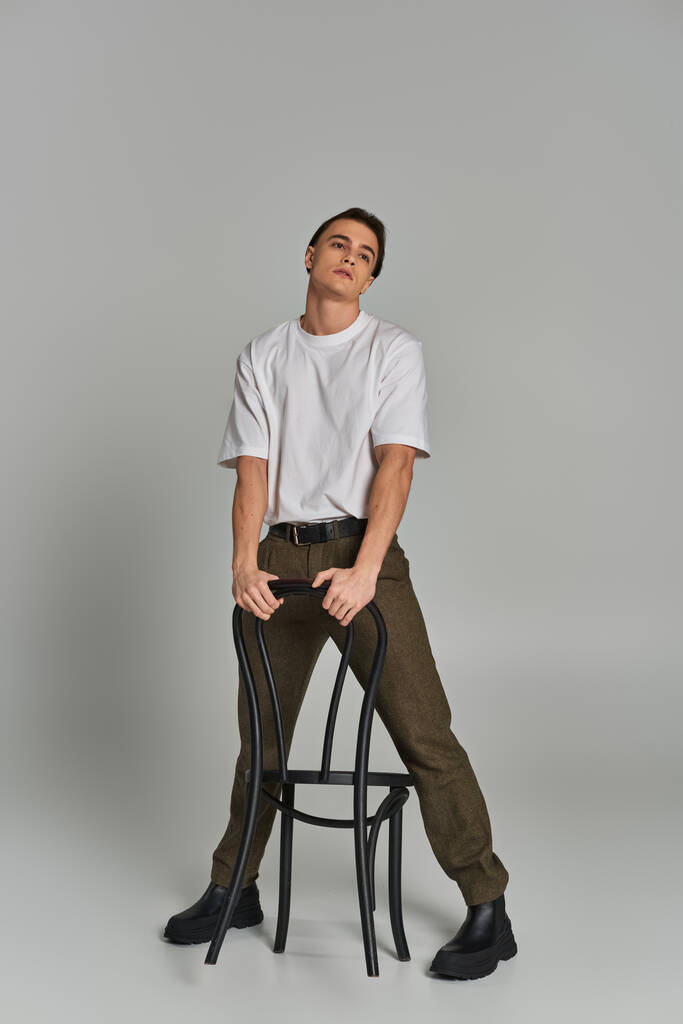 verleidelijke jongeman in trendy kleding zitten aantrekkelijk op stoel en weg te kijken op grijze achtergrond - Foto, afbeelding