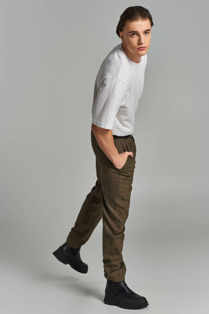 appealing young man in t shirt and brown pants posing attractively on gray backdrop and looking away - Photo, Image