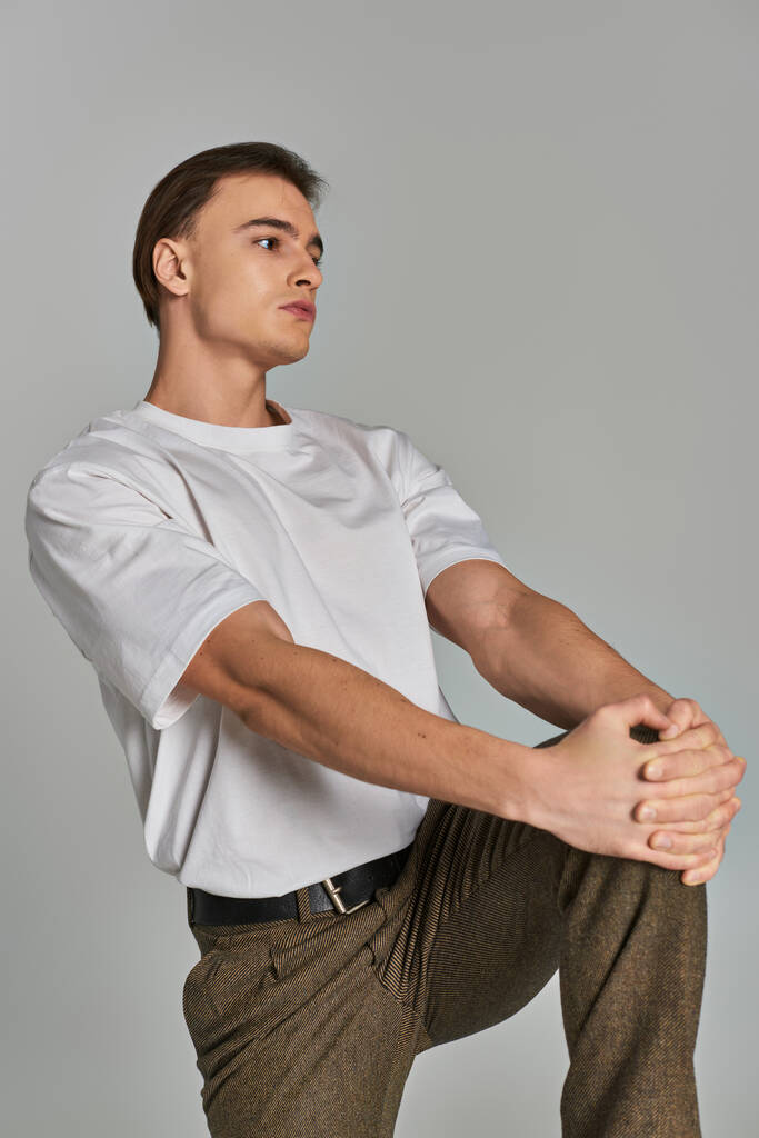 přitažlivý mladý muž v tričku a hnědé kalhoty pózuje atraktivně na šedém pozadí a dívá se jinam - Fotografie, Obrázek
