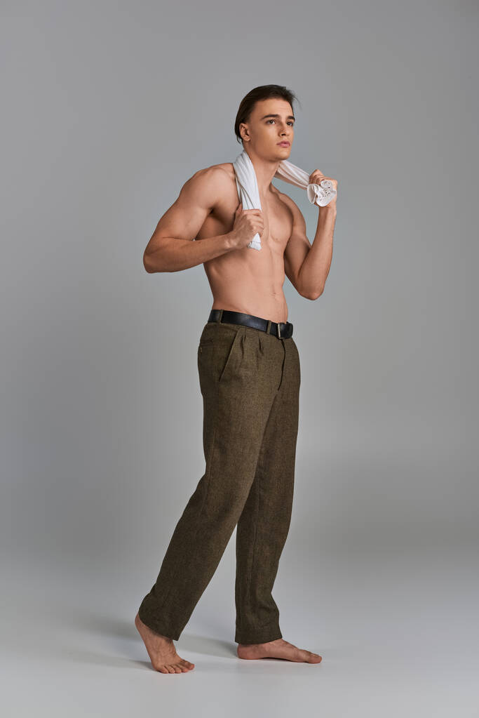 vonzó jóképű fiatalember barna nadrágban pózol félmeztelenül szürke háttér és félrenéz - Fotó, kép