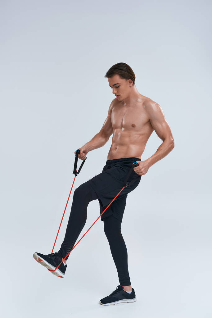 verleidelijke shirtloze sportieve jongeman in zwarte broek training met fitness expander en wegkijken - Foto, afbeelding