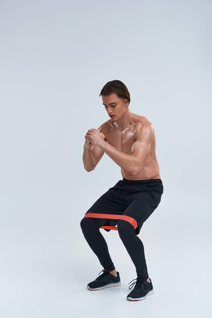 atletico sexy uomo senza maglietta in pantaloni neri utilizzando banda di resistenza per accovacciarsi su sfondo grigio - Foto, immagini