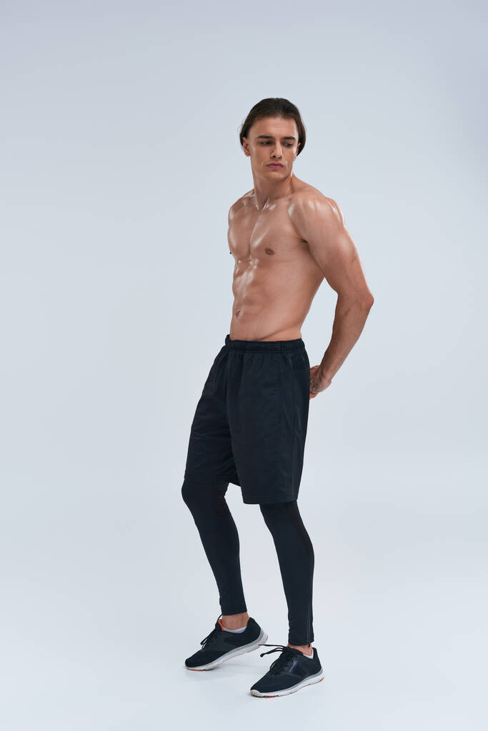 атлетичный красивый молодой человек позирует без рубашки в черных штанах и смотрит в сторону на сером фоне - Фото, изображение