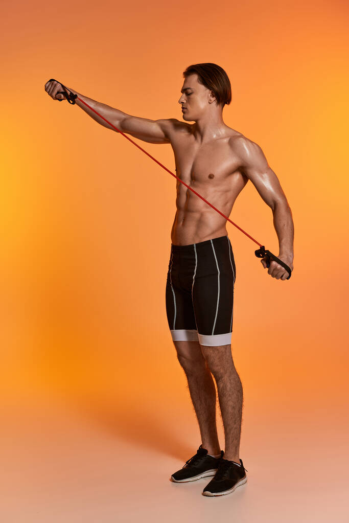 atractivo joven deportista en pantalones cortos negros posando en topless y haciendo ejercicio con expansor de fitness - Foto, Imagen