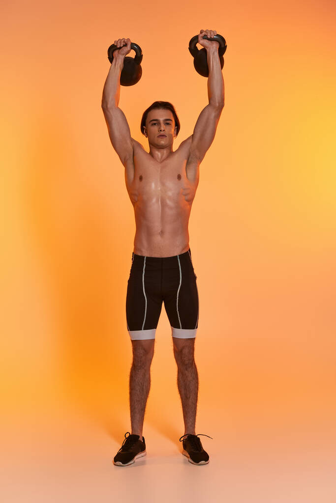 aantrekkelijk sportief shirtloos man oefenen met kettlebells en kijken naar de camera op levendige achtergrond - Foto, afbeelding