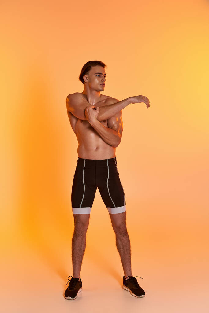 hombre guapo sin camisa en pantalones cortos negros haciendo ejercicio activamente y mirando hacia otro lado en el fondo naranja - Foto, imagen