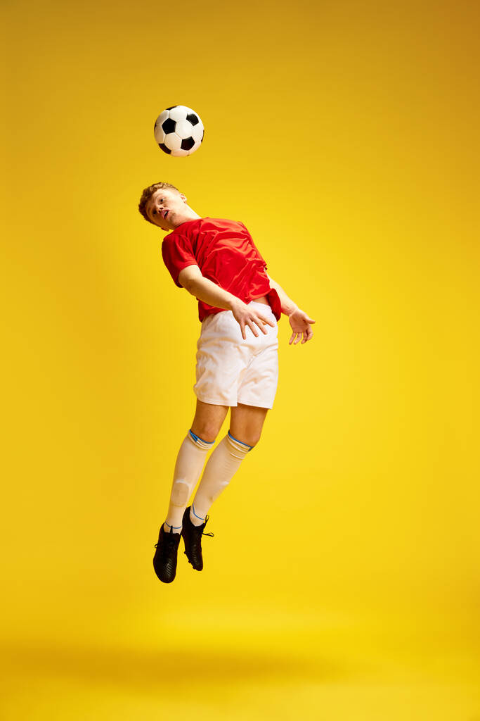 Obraz mladého muže v uniformě, fotbalista, nadšenec v pohybu, trénink, odpalování míče hlavou přes žluté pozadí studia. Koncepce aktivního životního stylu, mládeže, koníčků a emocí - Fotografie, Obrázek