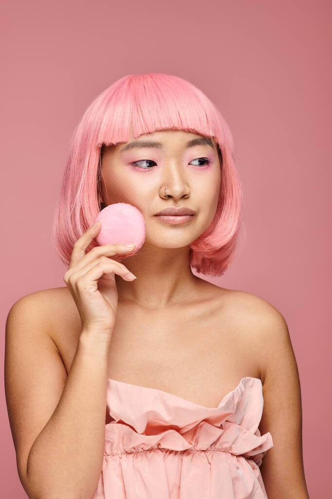 υπέροχο ασιατικό νεαρό κορίτσι με ροζ μαλλιά κοιτάζοντας προς τα πλάγια και κρατώντας να macaroon σε ζωντανό φόντο - Φωτογραφία, εικόνα