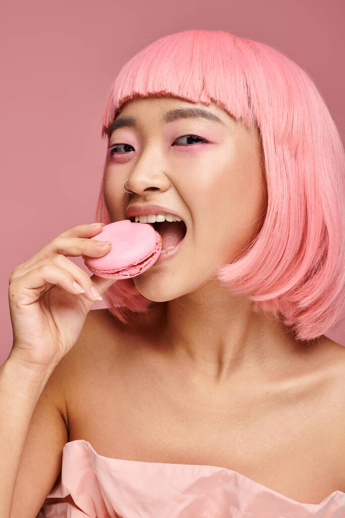 грайлива азіатська молода жінка з рожевим волоссям посміхається і їсть макаруни на яскравому фоні - Фото, зображення