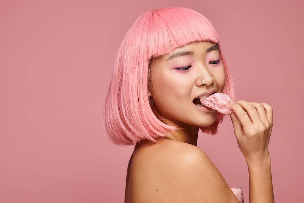 urocza azjatycka kobieta po dwudziestce z różowymi włosami jedząca mochi przez ramię na tętniącym życiem tle - Zdjęcie, obraz