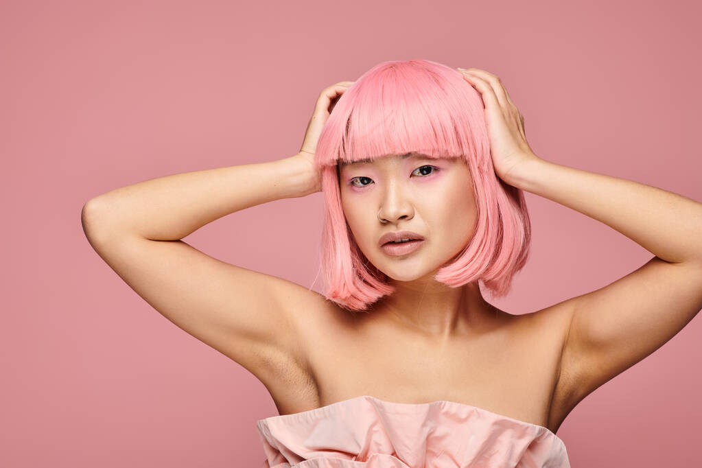 atractiva mujer asiática con pelo rosa y piercing posando con las manos en la cabeza sobre un fondo vibrante - Foto, imagen