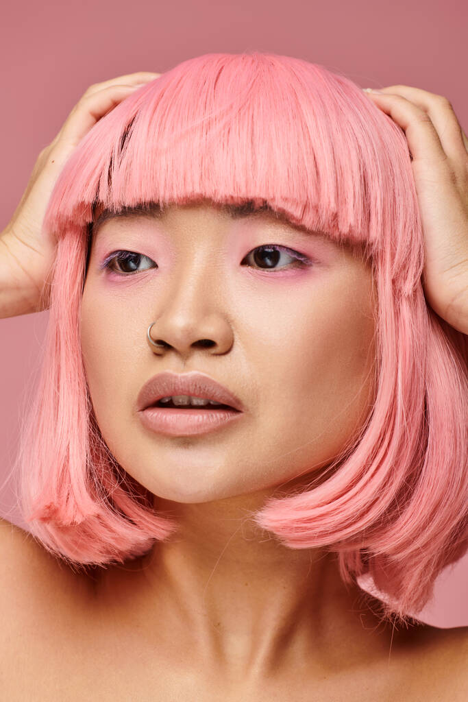 primer plano de mujer asiática con pelo rosa y piercing posando con las manos en la cabeza sobre un fondo vibrante - Foto, Imagen