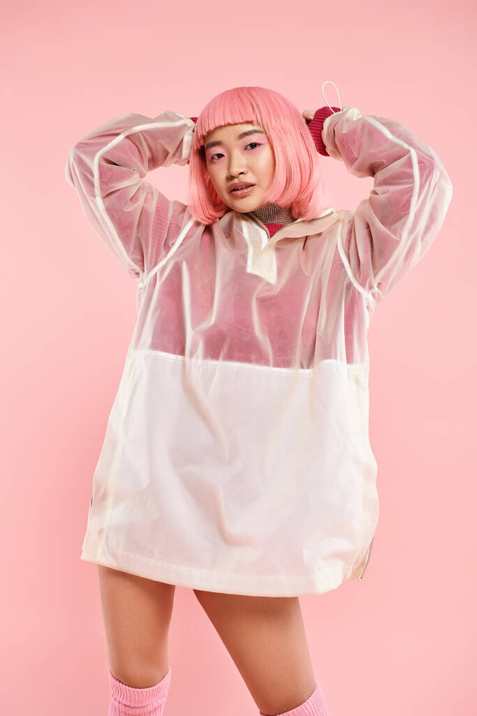 elegante sonriente asiático mujer en 20s con rosa pelo posando con las manos en la cabeza en vibrante fondo - Foto, Imagen