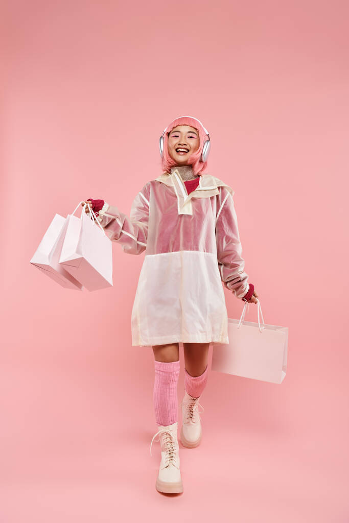 活気に満ちた背景を歩くヘッドフォンでピンクの髪とショッピングバッグと幸せなアジアの少女 - 写真・画像