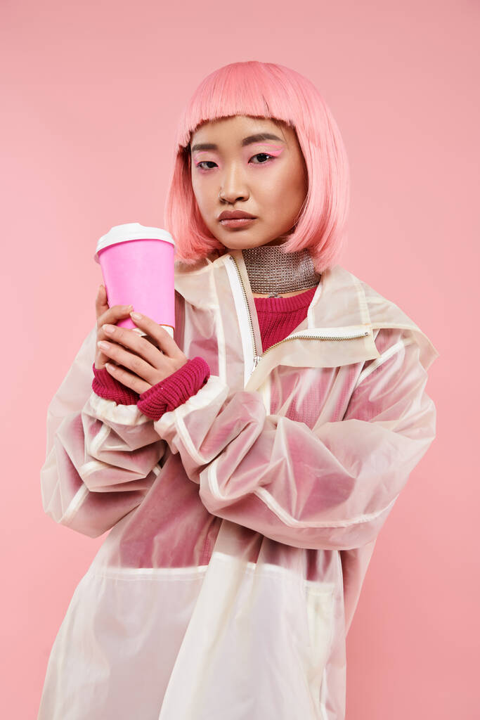 με αυτοπεποίθηση ασιατική νεαρή γυναίκα με ροζ μαλλιά ποζάρουν με φλιτζάνι καφέ σε ζωντανό φόντο - Φωτογραφία, εικόνα