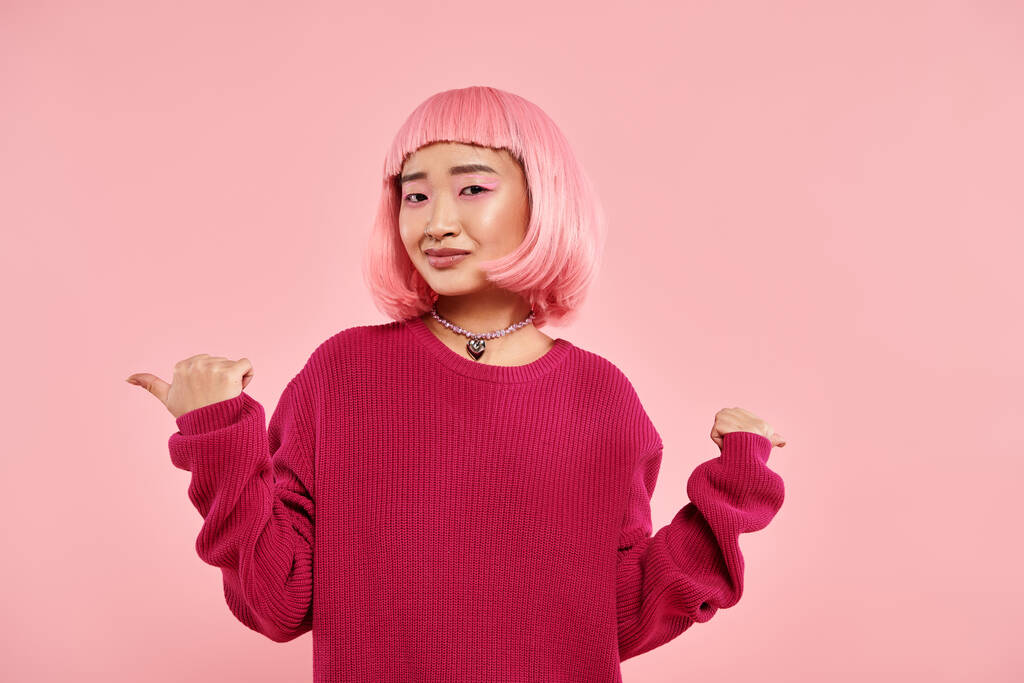 young woman in vibrant jumper and pink hair pointing away with her thumbs and looking at camera - Photo, Image