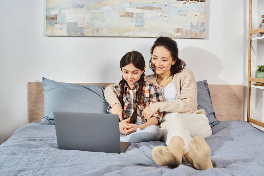 Μια μητέρα και η κόρη κάθονται κοντά σε ένα κρεβάτι, επικεντρώθηκε σε μια οθόνη laptop με ενδιαφέρον και σύνδεση. - Φωτογραφία, εικόνα